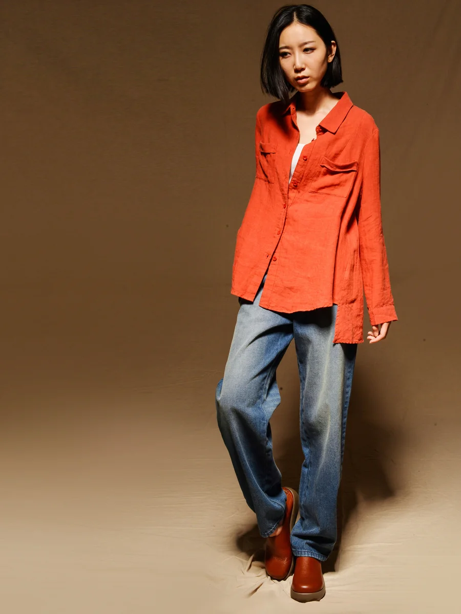 Model in a rust-orange shirt and mid-blue straight jeans against a brown studio backdrop
